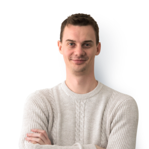 A clean shaven man with short brown hair, wearing a cream jumper with arms folded